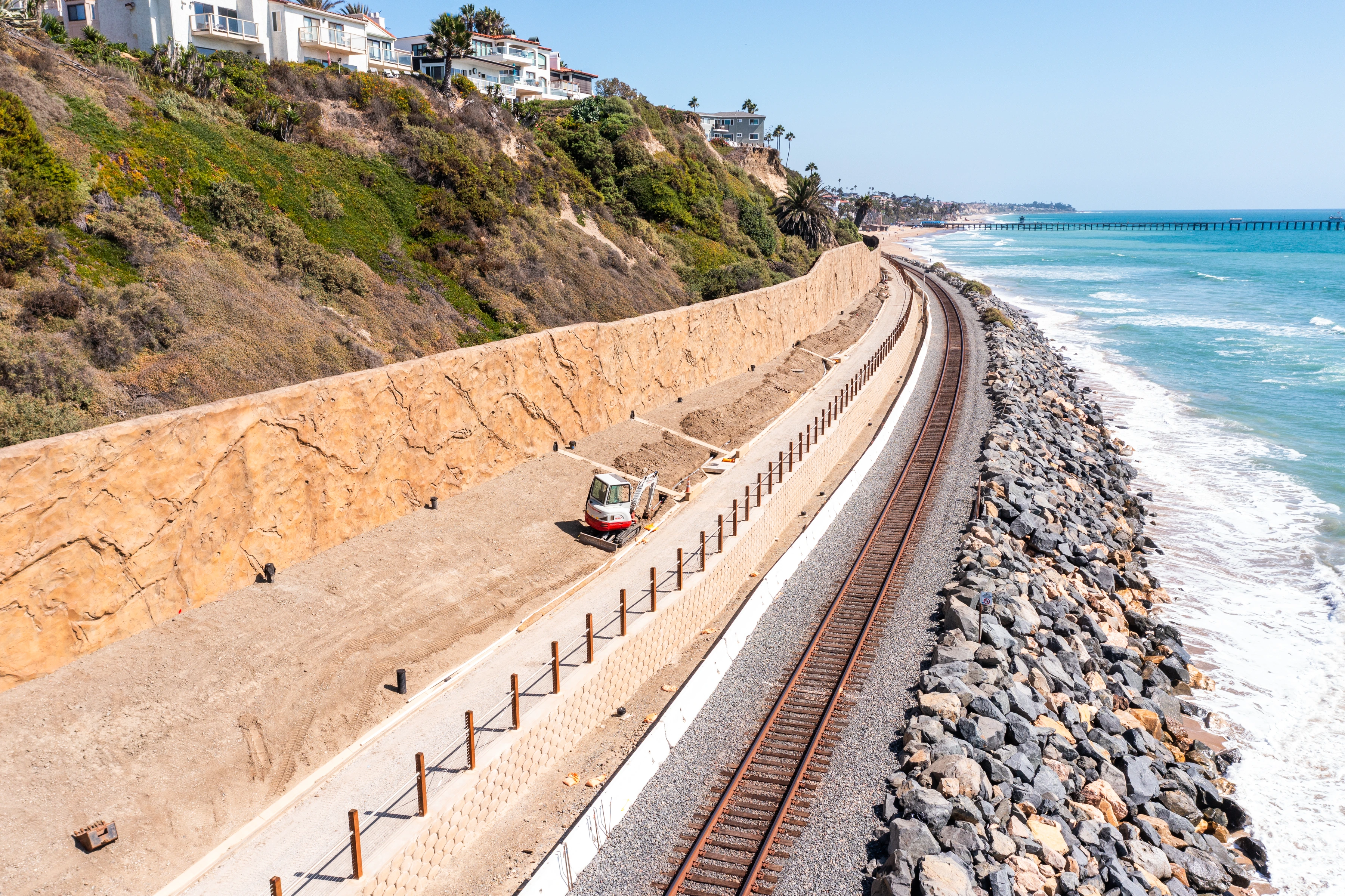 Elevated view of railroad tracks curving between a graded bluff-side work area and the rock-armored shoreline at Mariposa Point.