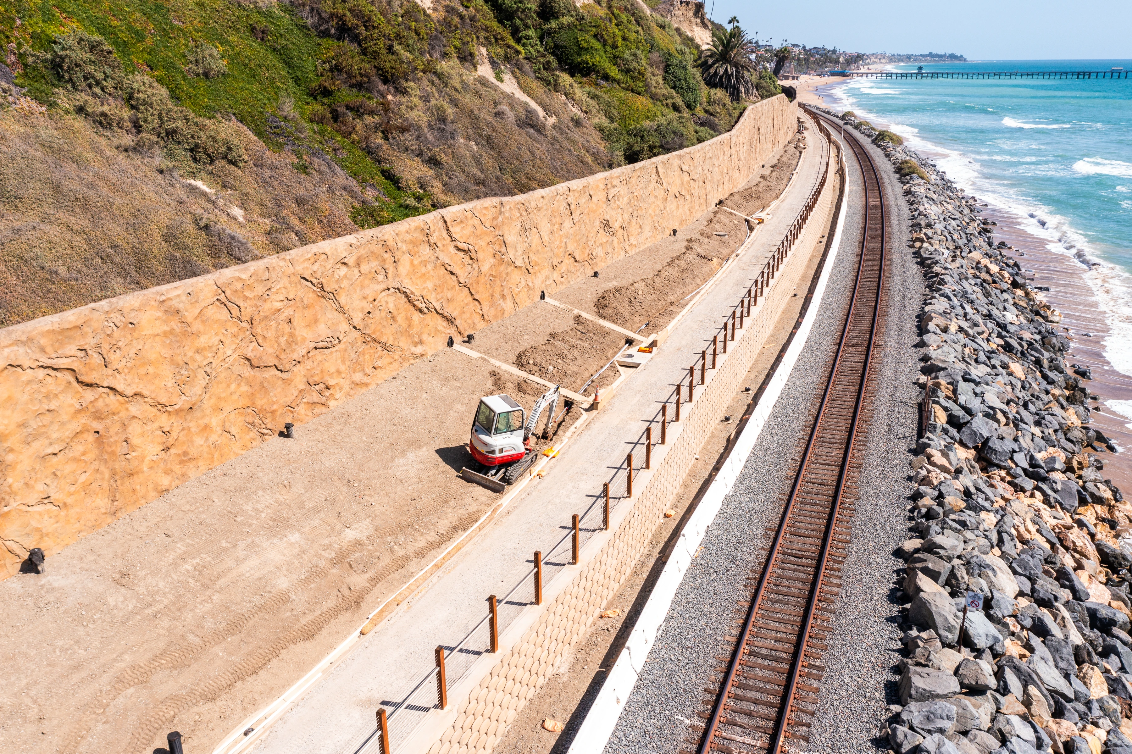 Detailed view of a coastal rail work area with a compact excavator, shallow excavation, patterned lower wall, fencing components, and staged materials.