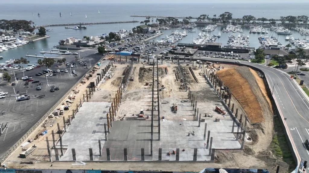 Wide view of the Dana Point Harbor site with poured concrete areas, excavated earth, upright structural elements, staged materials, compact equipment, and a marina in the background.