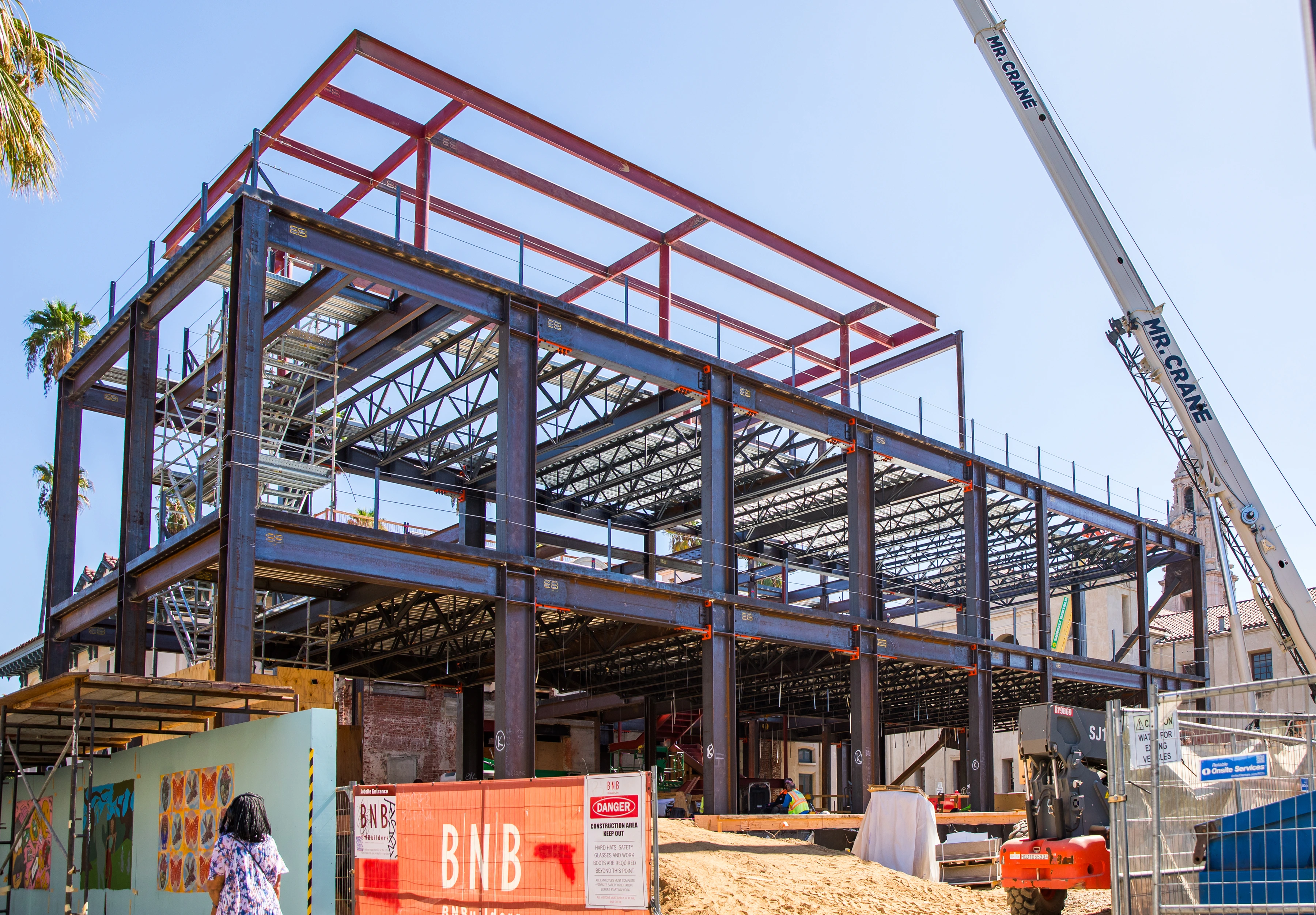 Wide view of a multilevel steel frame with open floor bays, temporary stairs, fencing, and a crane boom.
