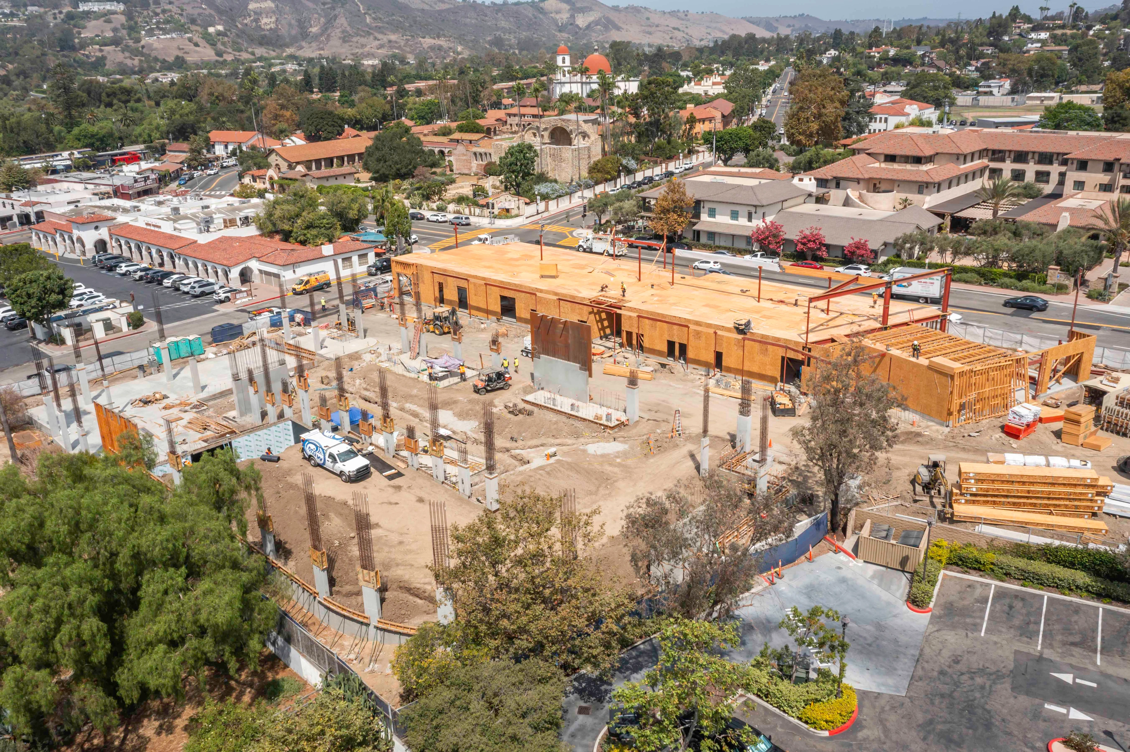 Wide construction view with concrete columns in the foreground, a wood roof deck beyond, and staged materials and equipment across the site.